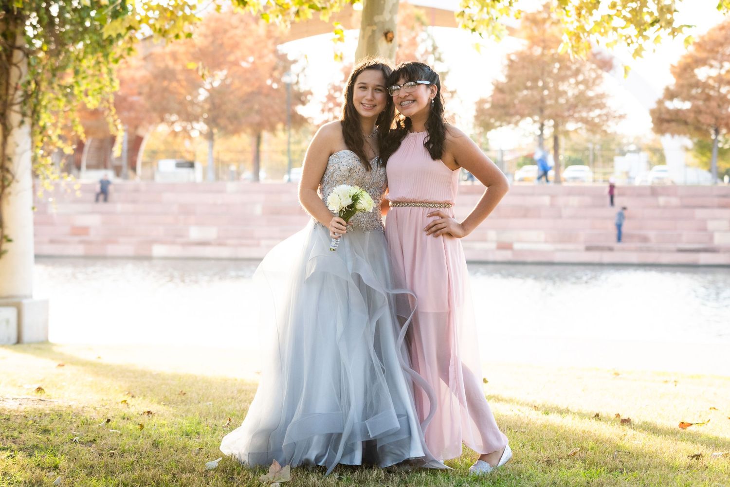 Two formal attendants pose together in complementary blue and pink gowns outdoors.