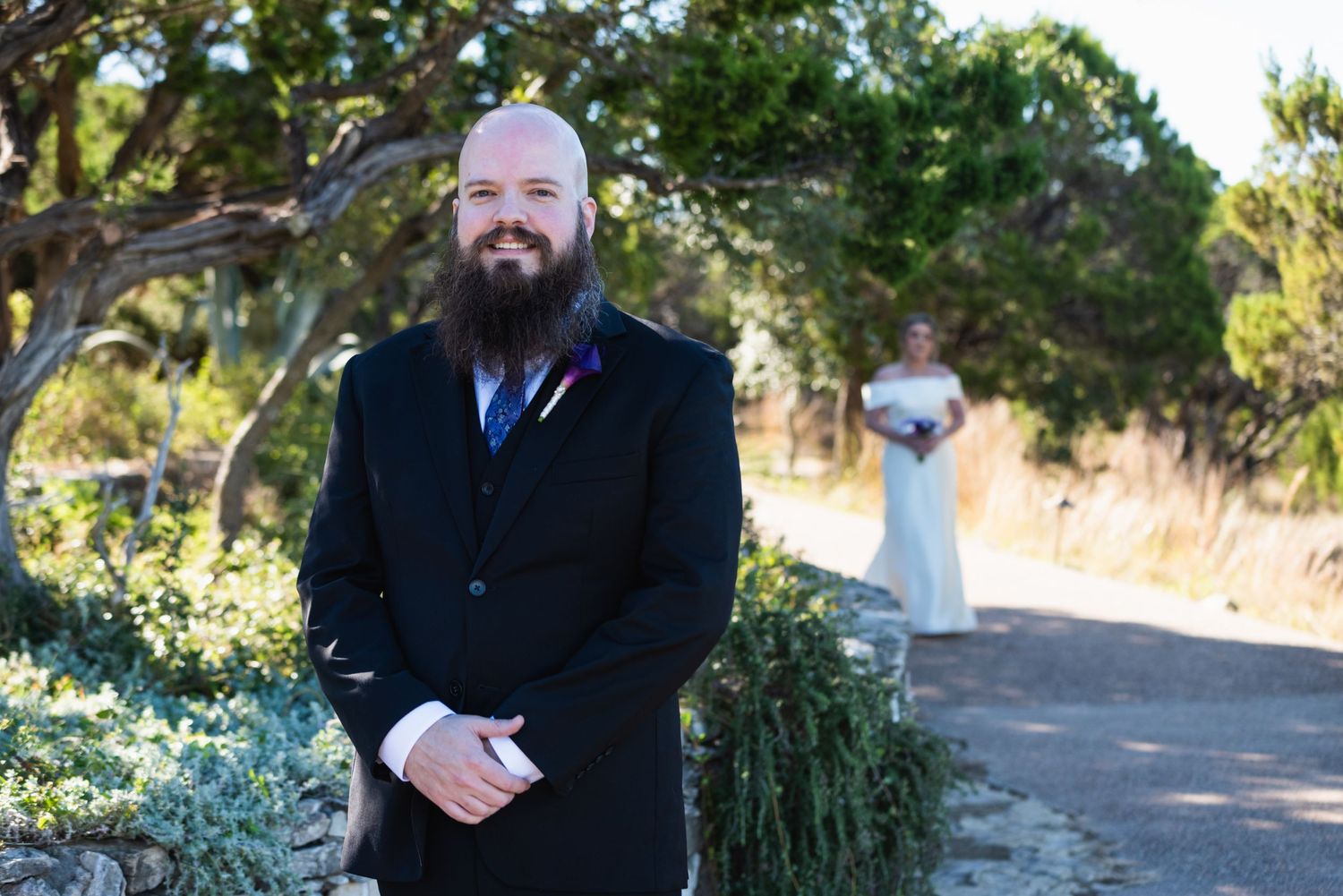 Person in dark suit stands outdoors with blurred figure in white dress in background.