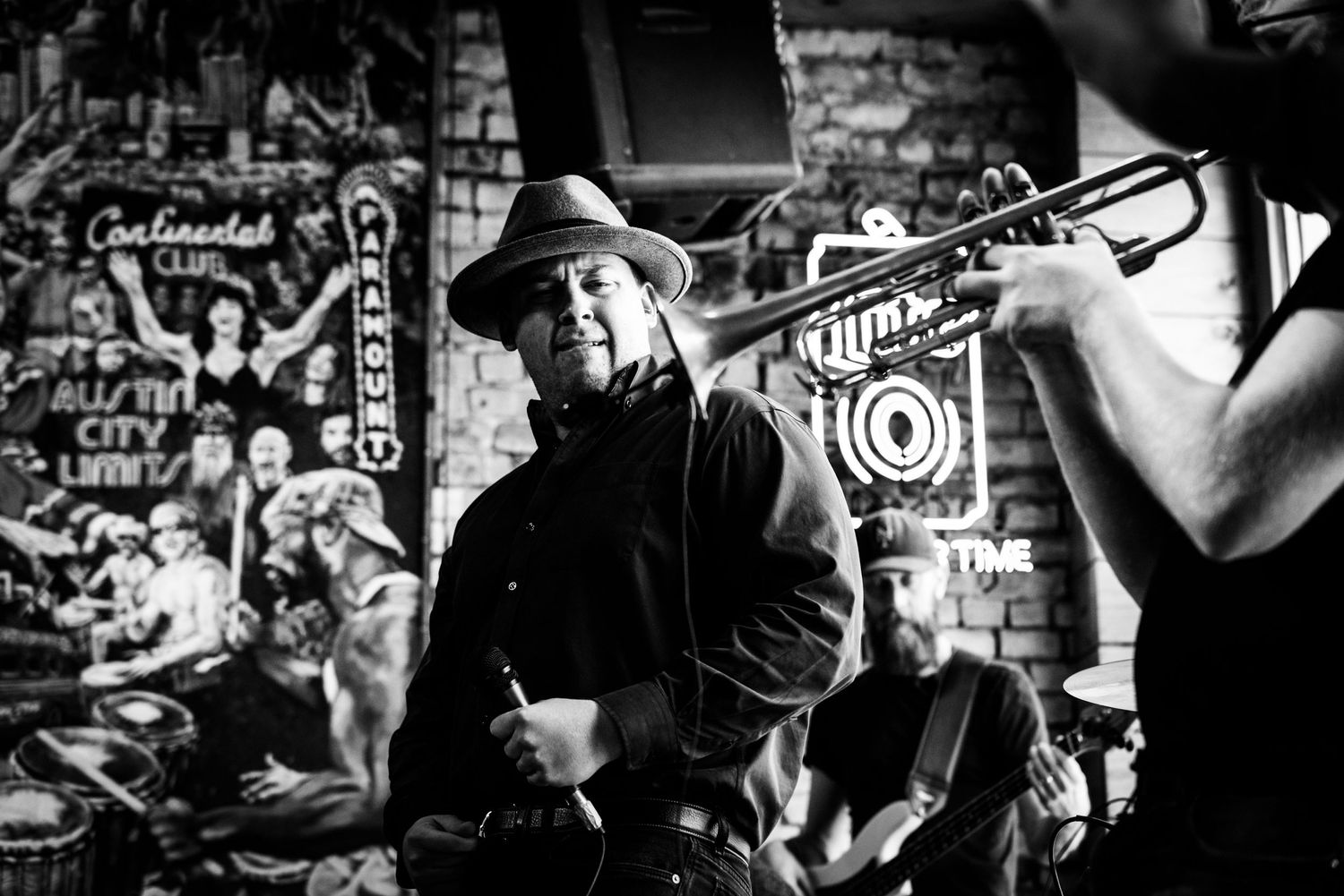Black and white photograph of a live music performance in a bar with a brick wall backdrop and neon signs.