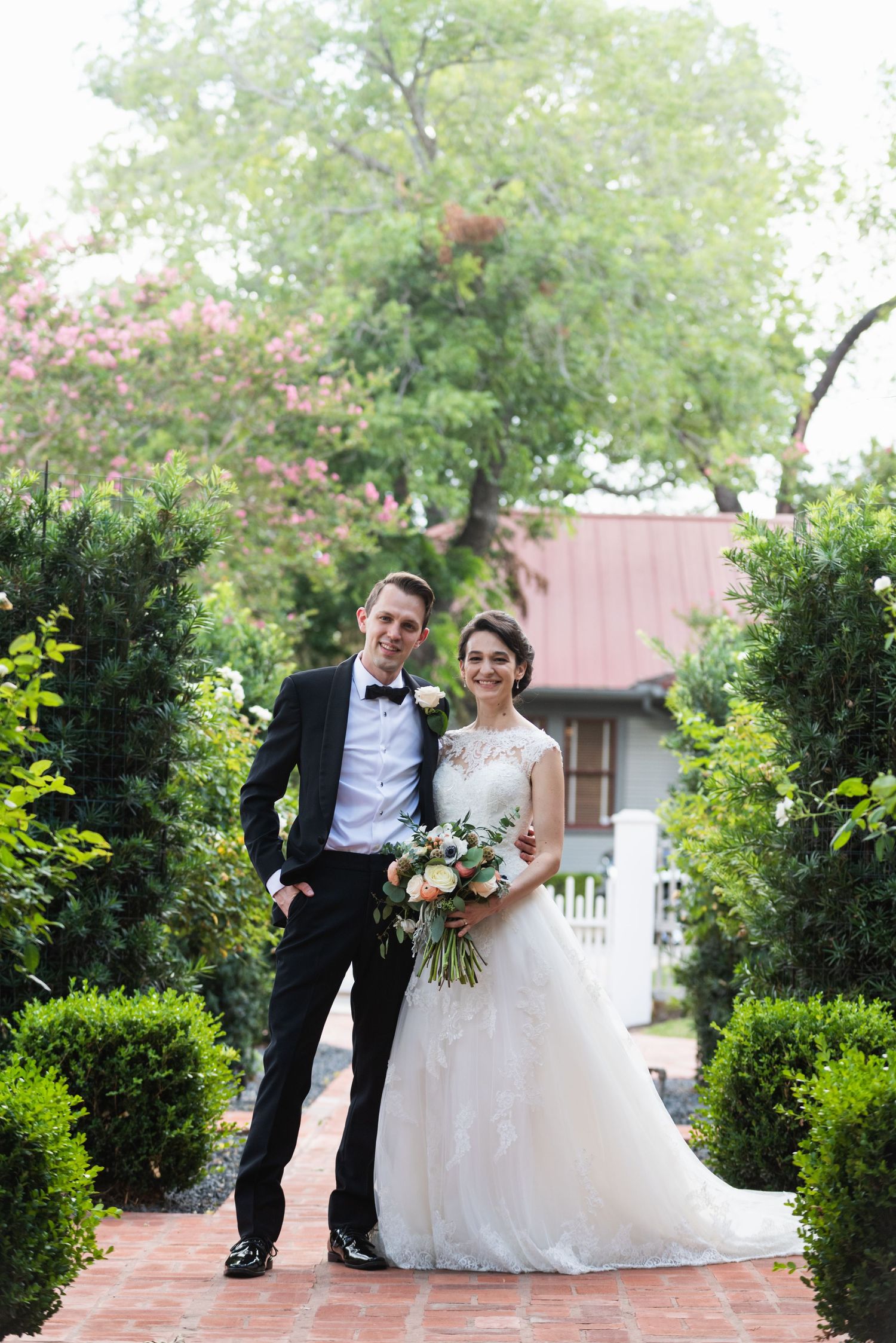 A classic wedding portrait on a brick pathway surrounded by manicured gardens and greenery.
