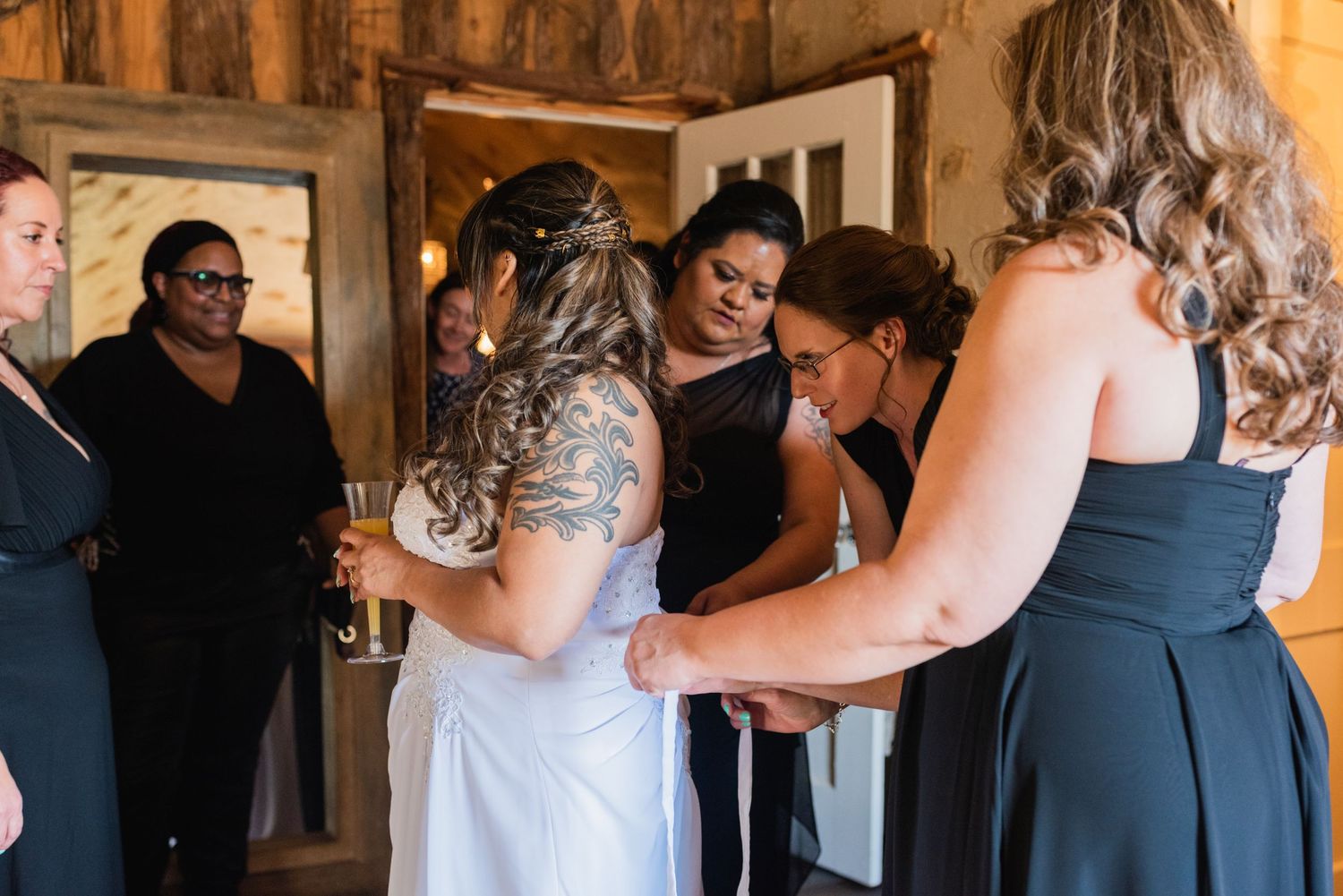Bridesmaids in navy dresses help adjust the bride's gown in a wooden interior.