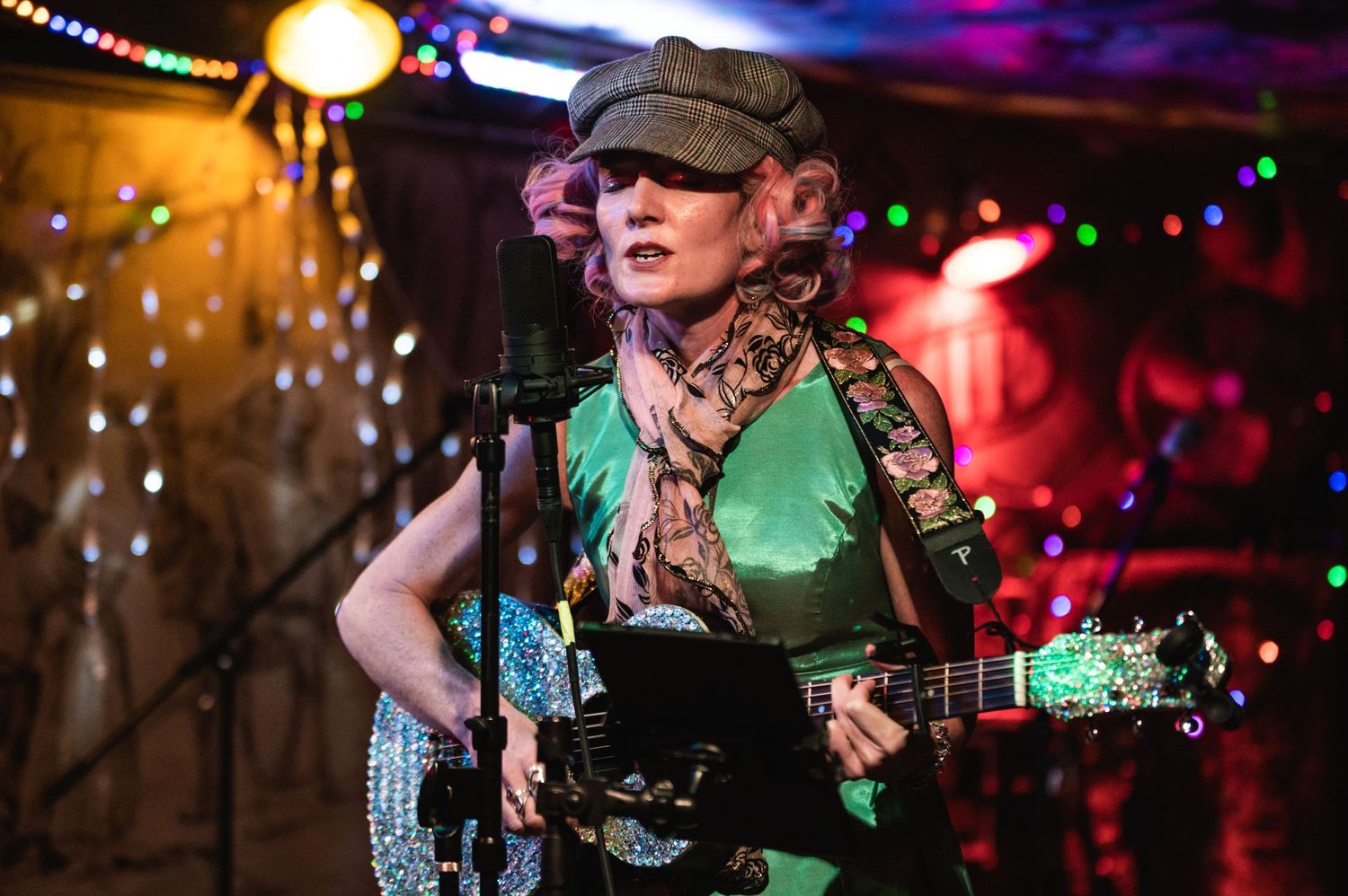 Musician performs with guitar under colorful stage lighting with string lights in background.