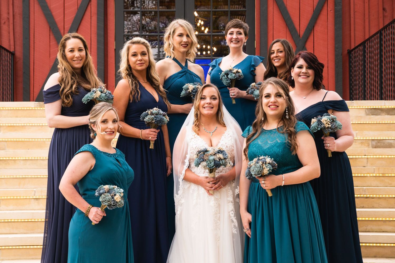 A bridal party in navy and teal dresses poses on stone stairs with bouquets.