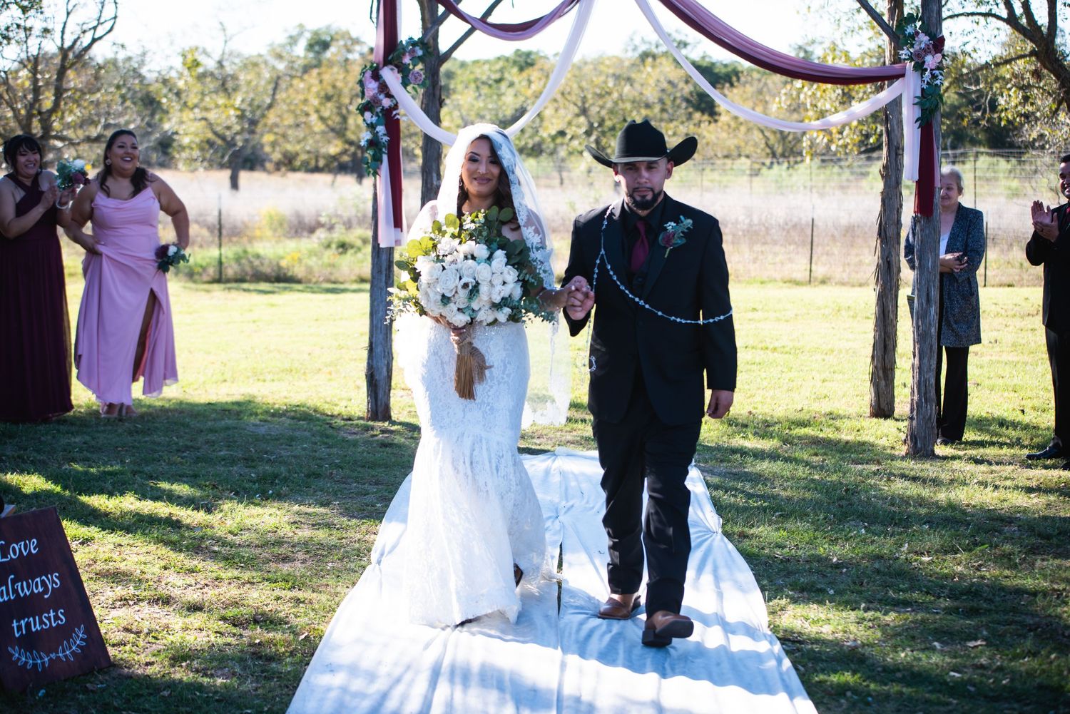 Newlyweds walk down white aisle runner under purple-draped wooden arch at outdoor ceremony.