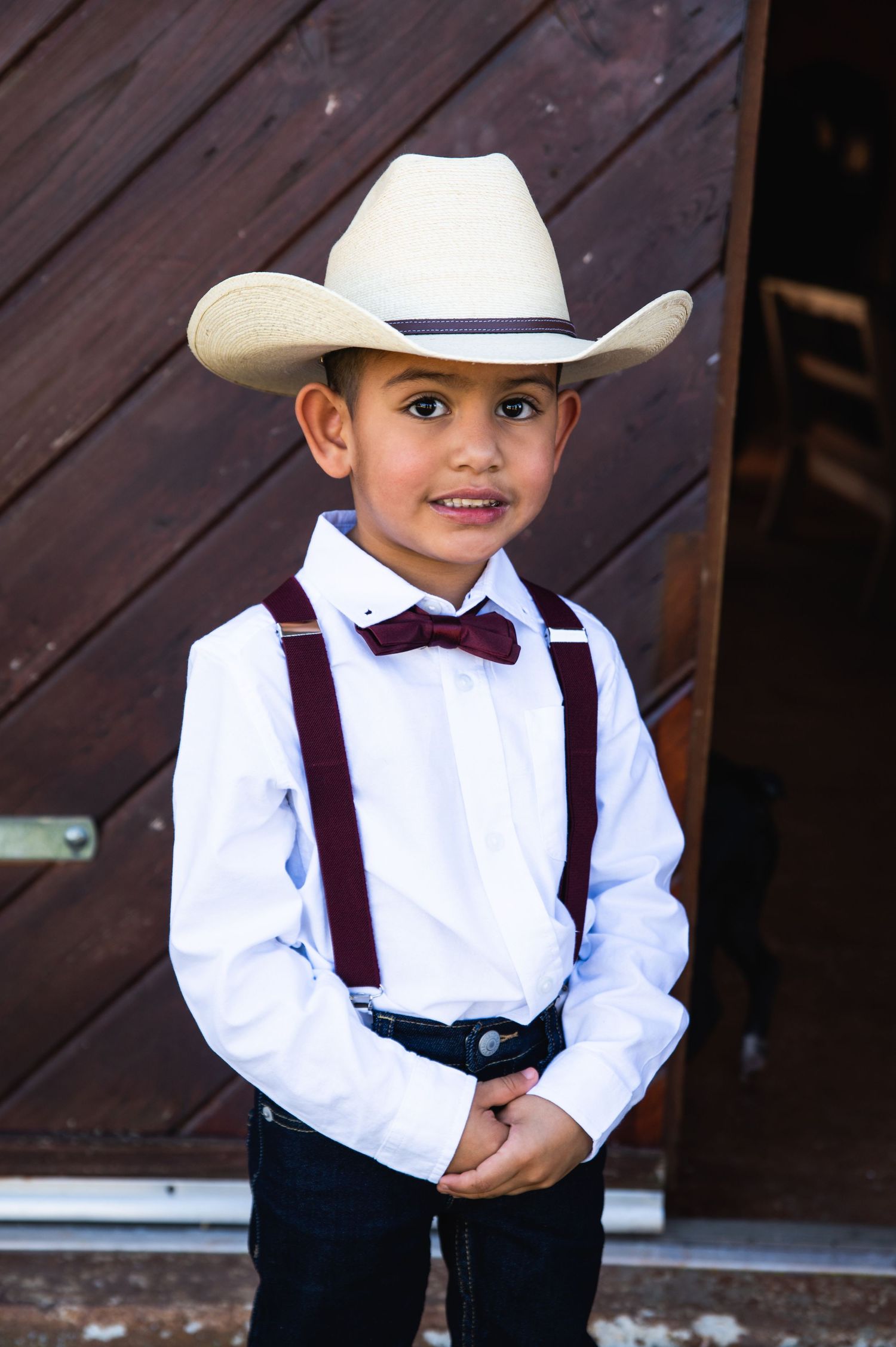 Young ring bearer in white cowboy hat, bow tie and suspenders smiles at camera.