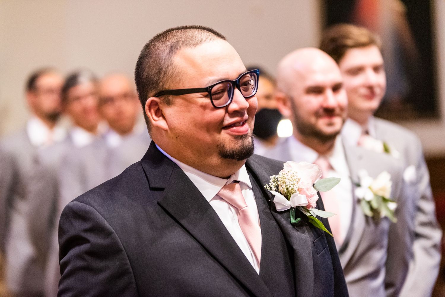Groom in black suit and pink tie smiles during wedding ceremony.