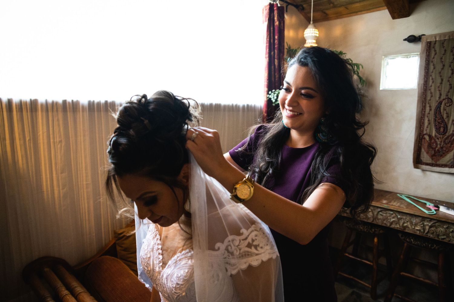 A helper adjusts a delicate wedding veil in warm indoor lighting.