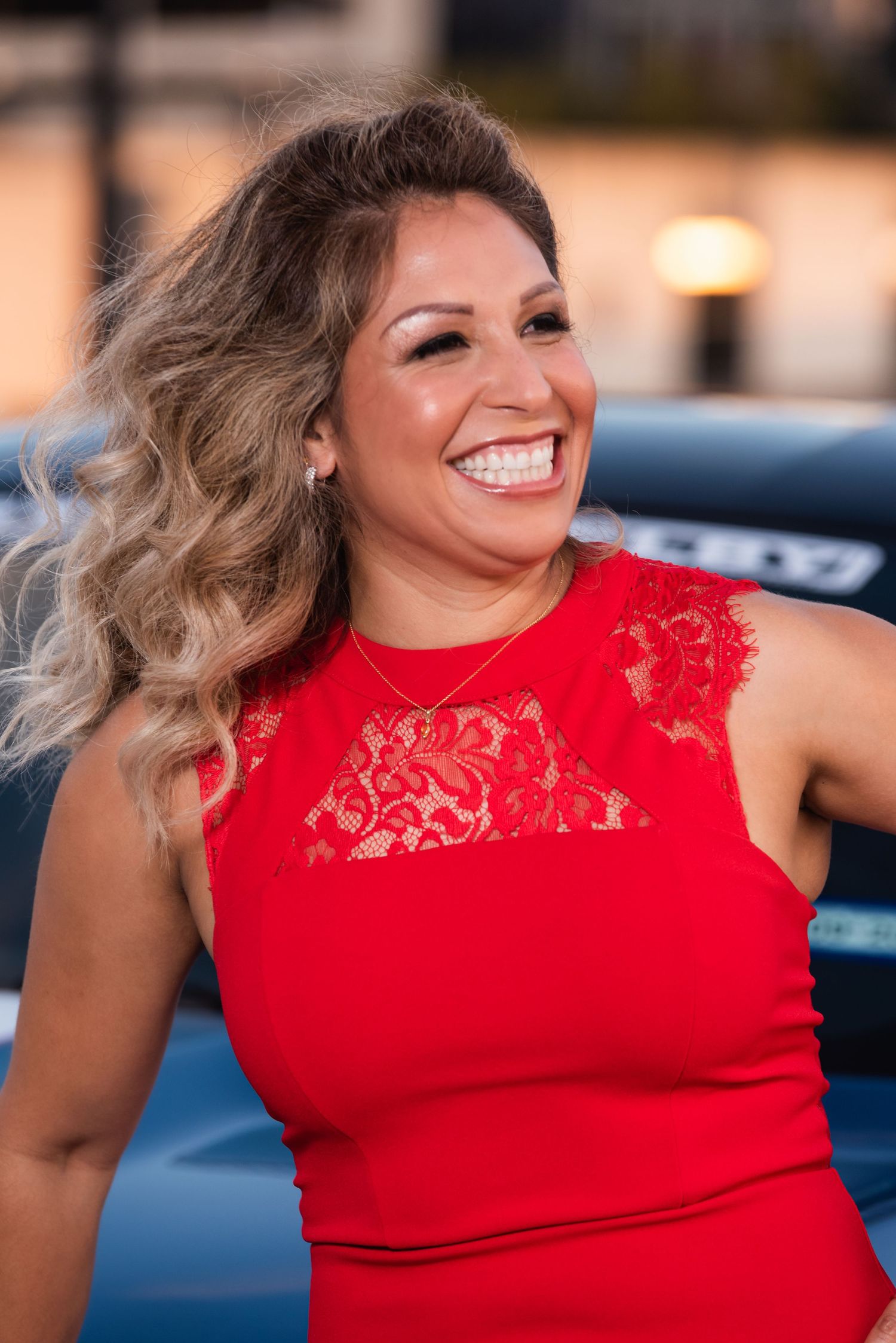 Portrait against blue sports car featuring red lace dress and flowing hair.