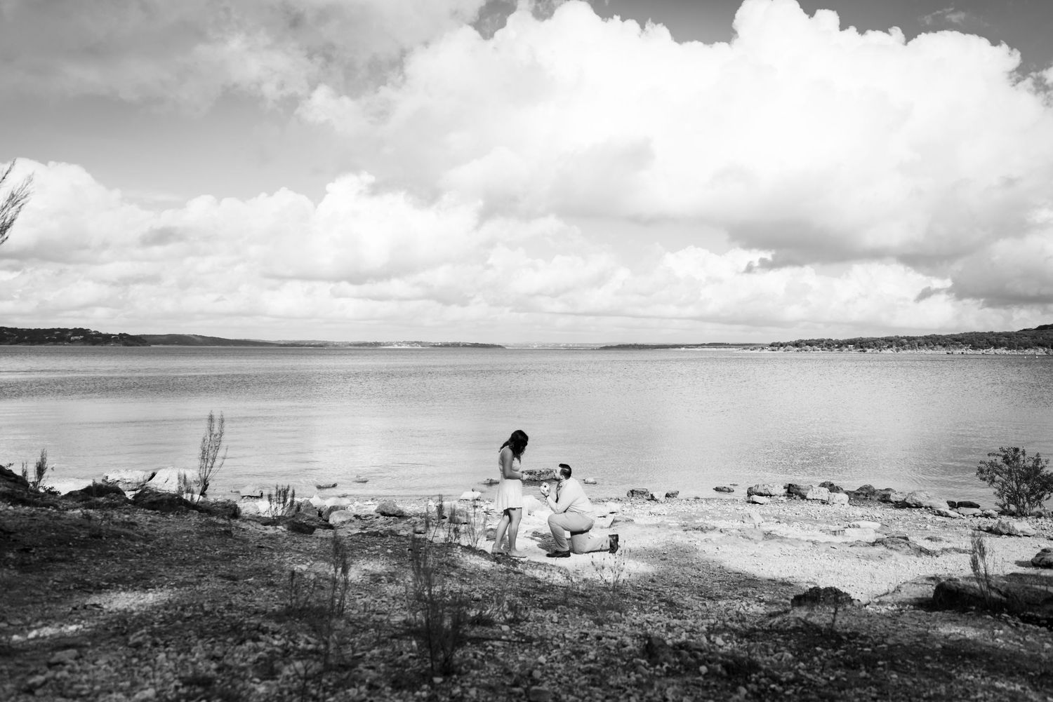 Black and white landscape photo of person sitting by lake with cloudy sky.