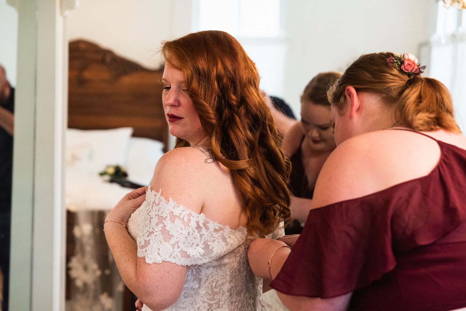A person in a white lace off-shoulder wedding dress being helped with preparations.