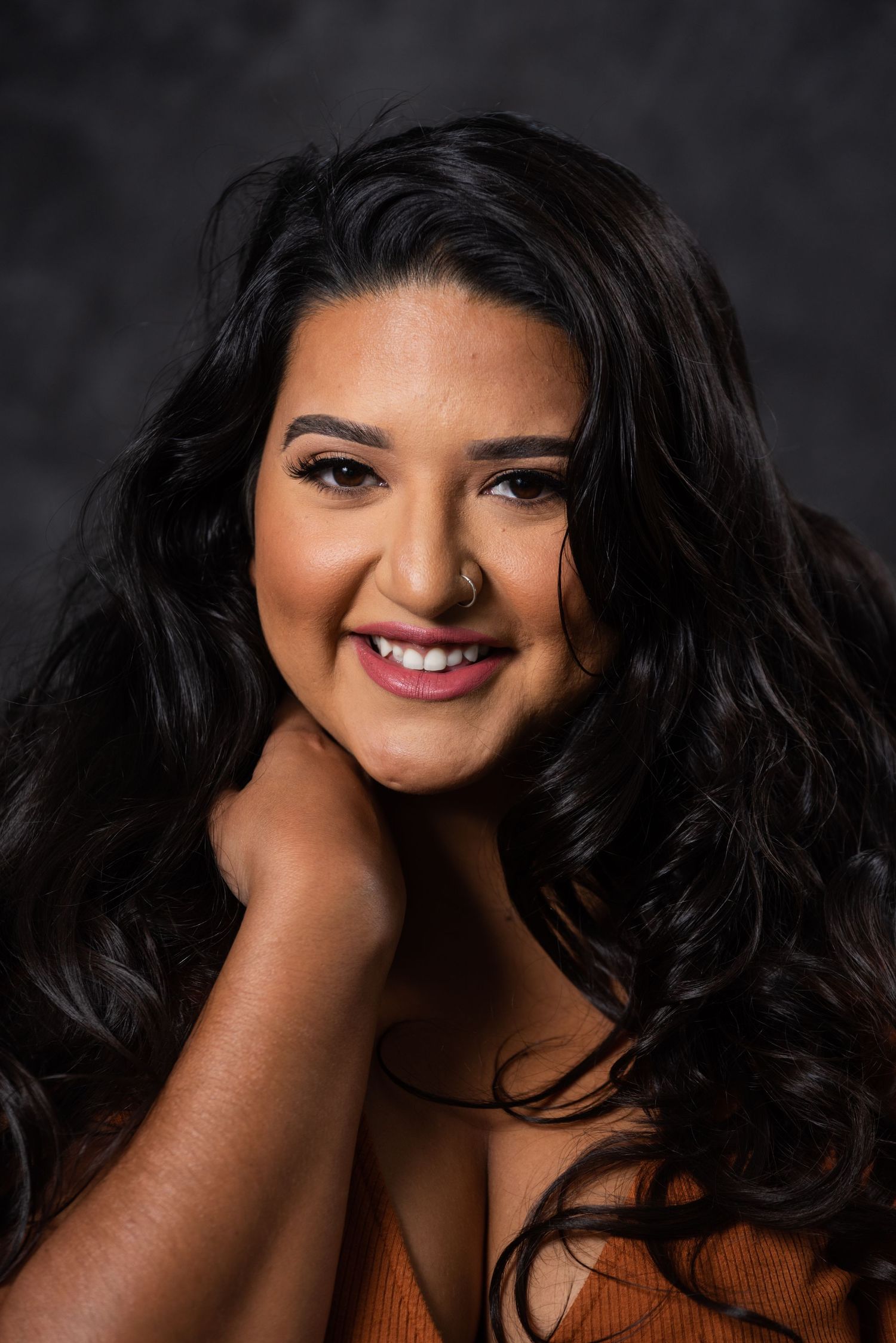 Portrait of woman with long dark hair and orange top smiling against dark background.