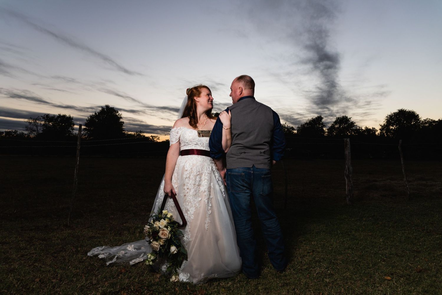 Newlyweds pose together against a sunset sky holding a floral bouquet.
