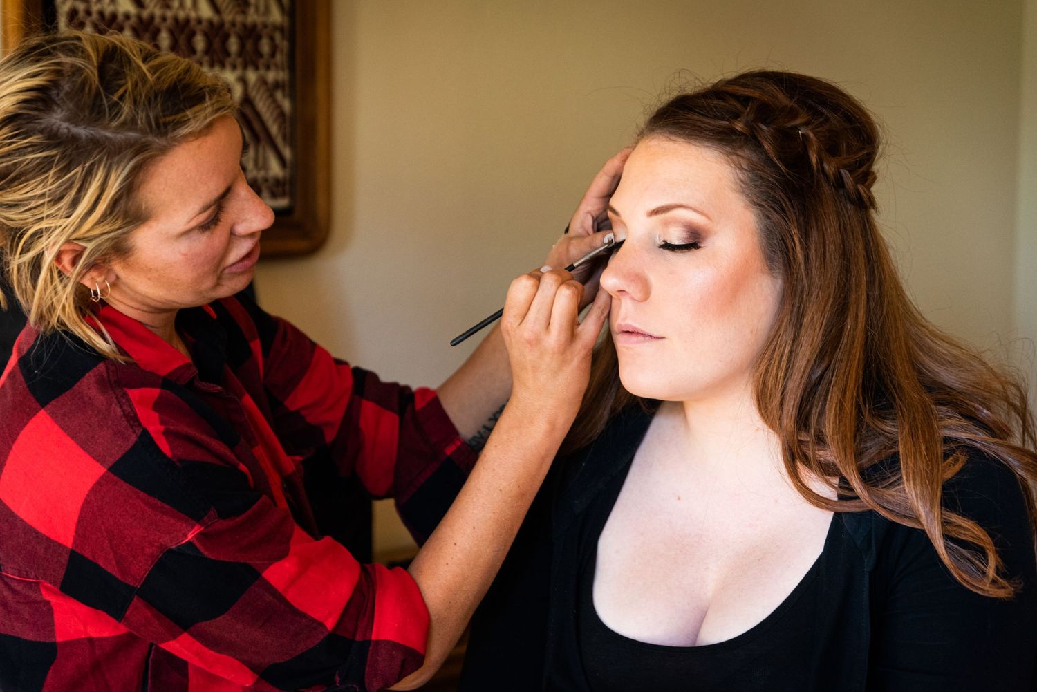 A makeup artist wearing a plaid shirt applies eye makeup to a client.