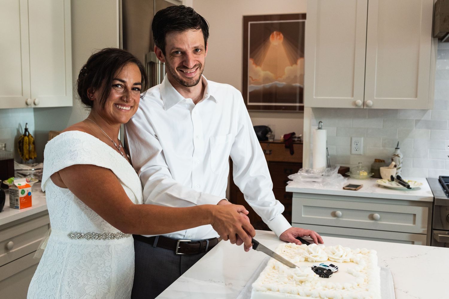 A couple cuts their wedding cake together in a modern white kitchen.