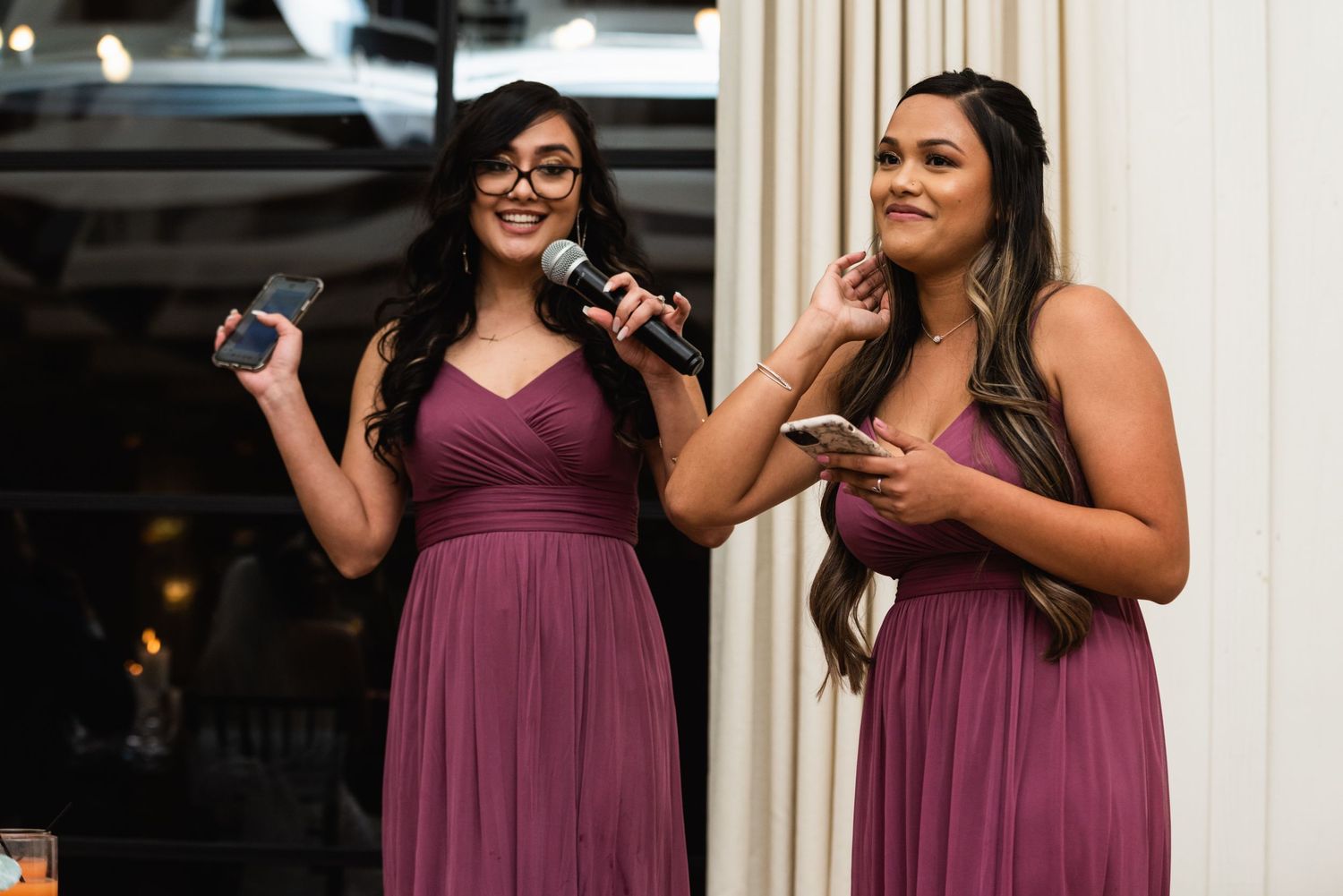 Two bridesmaids in burgundy dresses giving speeches with microphones at a wedding reception.