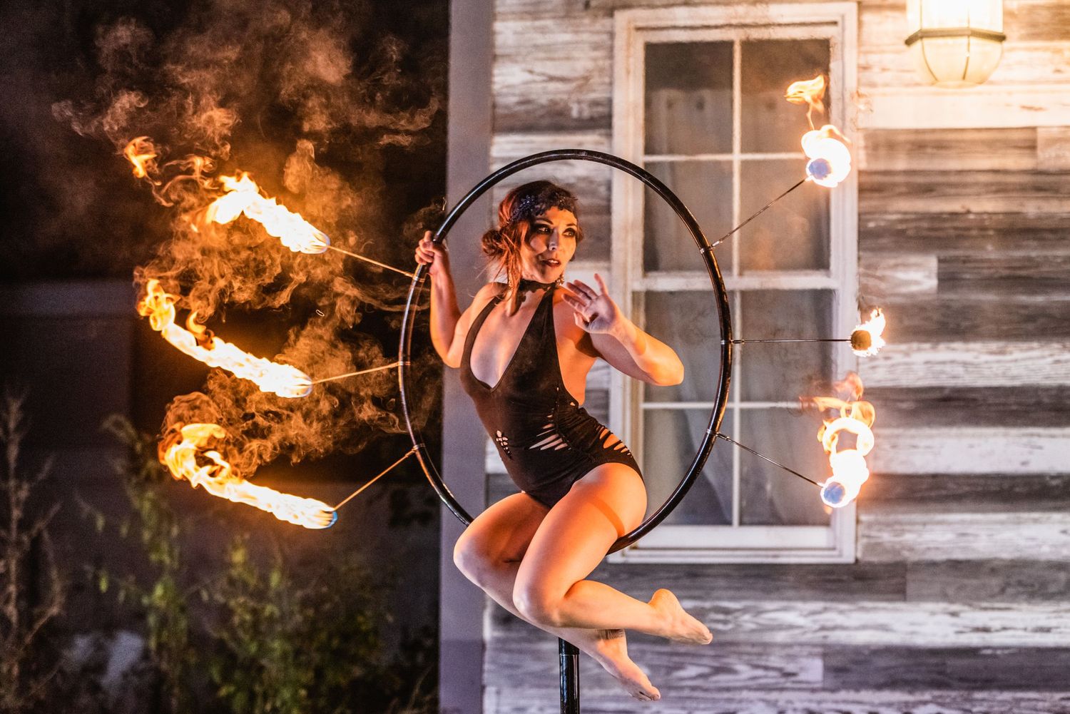 Performer on aerial hoop with fire elements against rustic building backdrop.