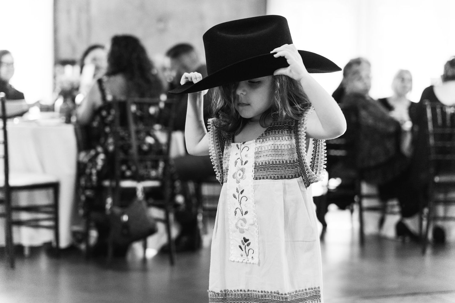 A small child wearing a hat and white dress plays during a wedding reception.