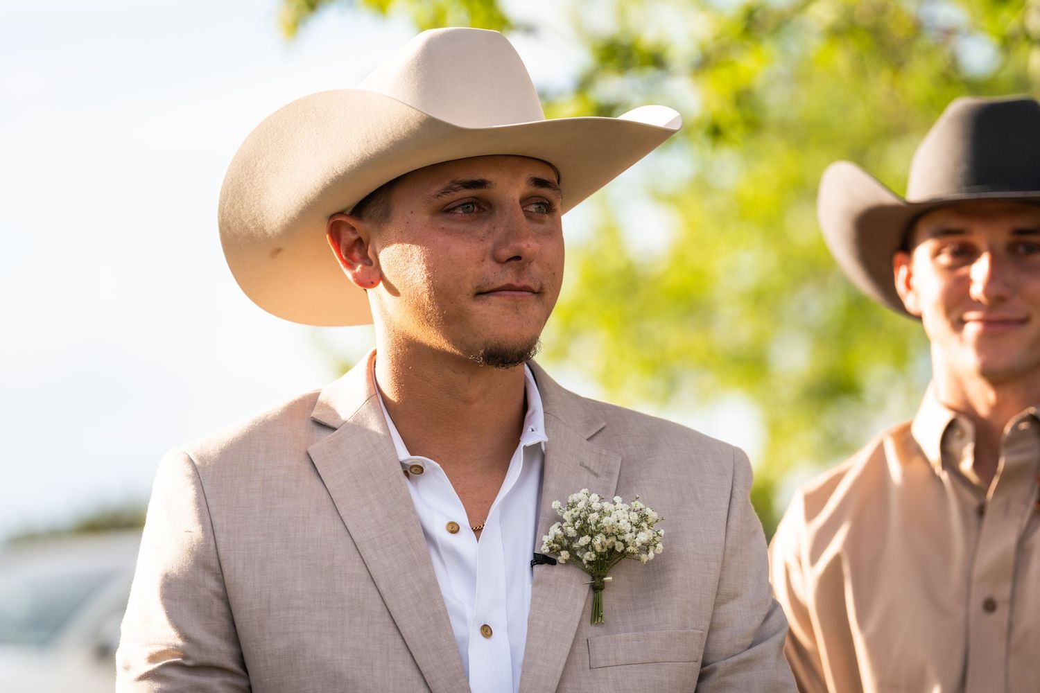 A groom in a beige suit and white cowboy hat smiles during an outdoor wedding.