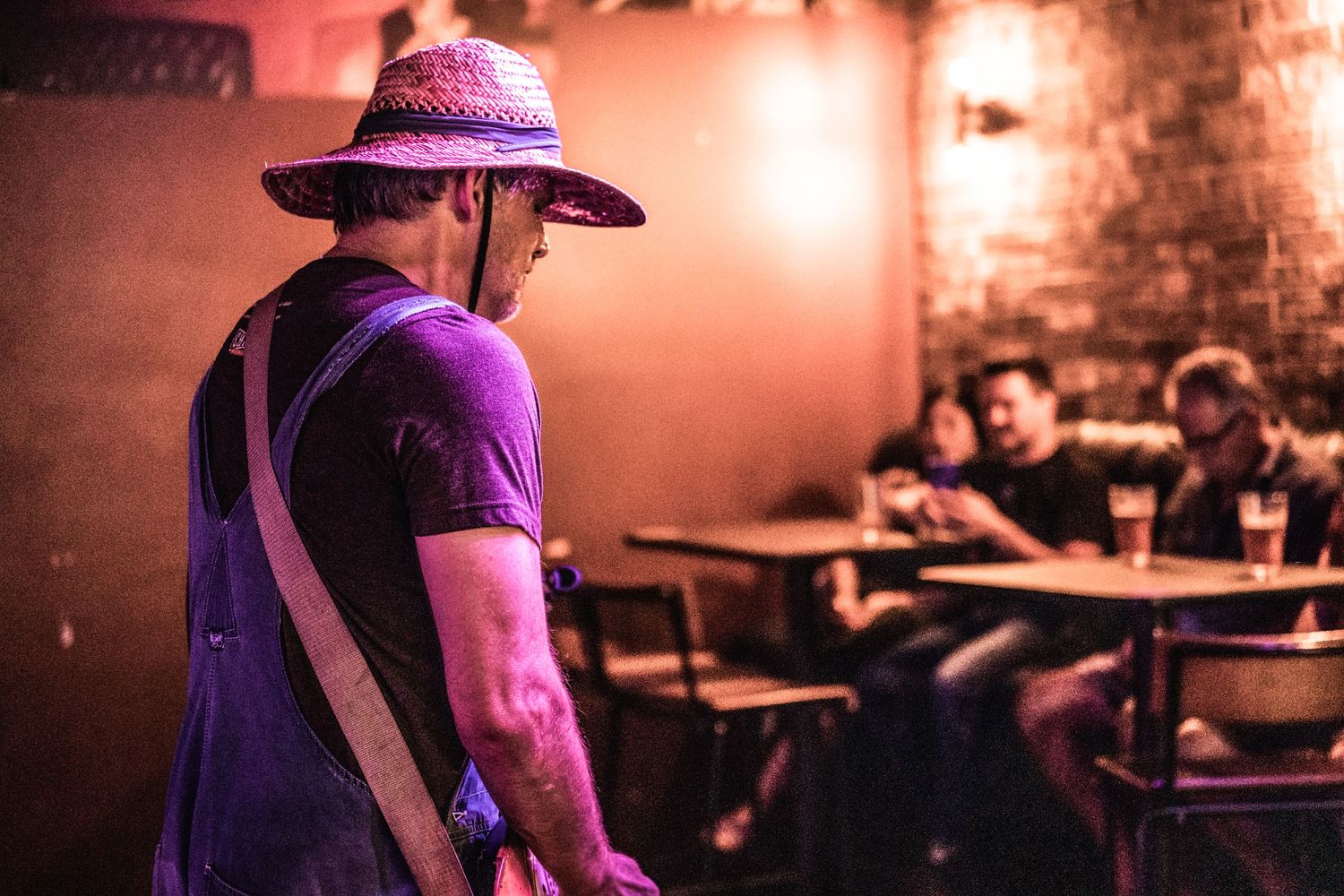 Musician performs in straw hat and overalls at intimate venue with brick walls.