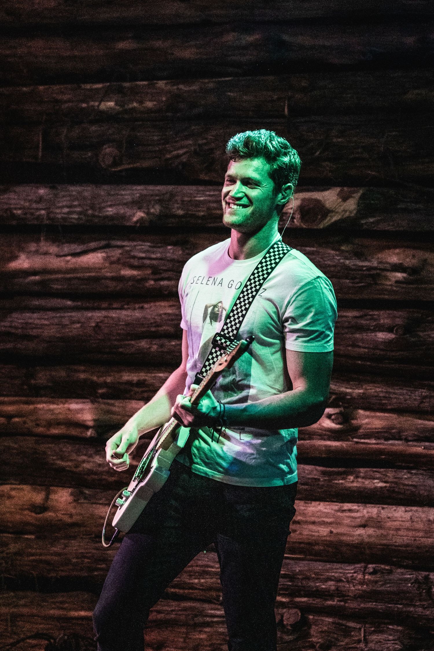 Guitarist in a white t-shirt performs enthusiastically against a rustic wooden backdrop under green lighting.