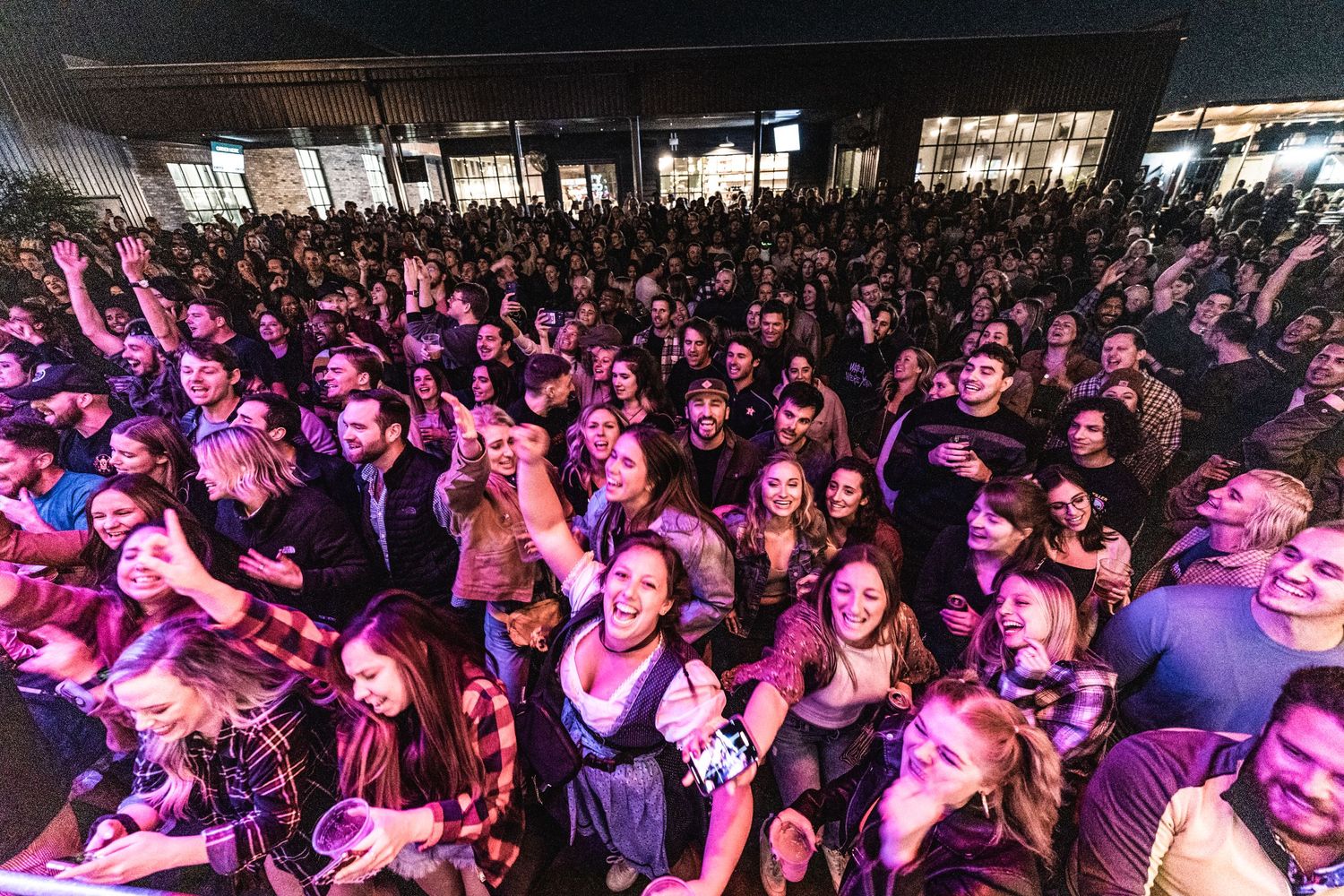Large concert crowd illuminated by pink and purple stage lights at outdoor venue.