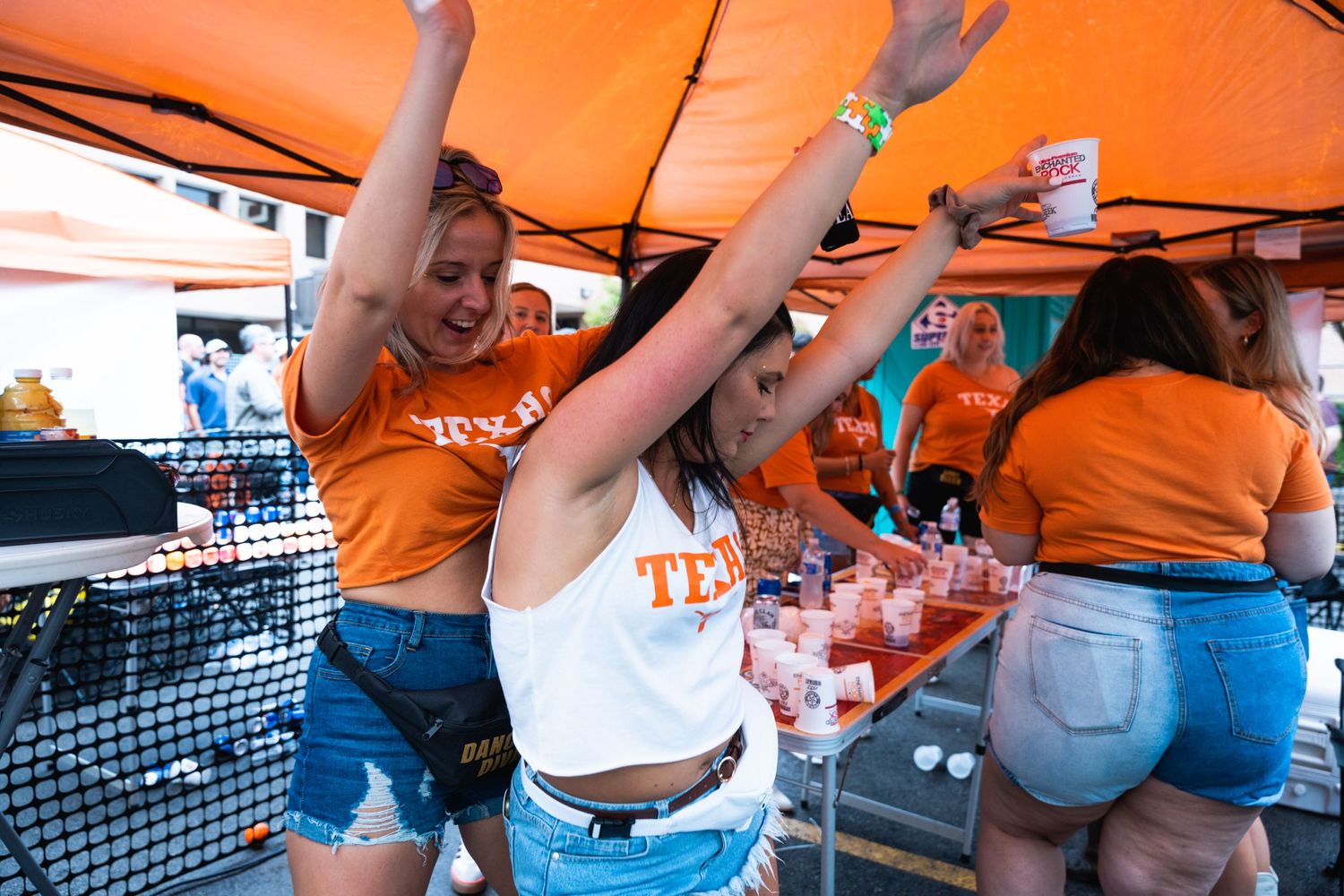 Energetic crowd celebrates under an orange tent with drinks and raised hands at an outdoor event.