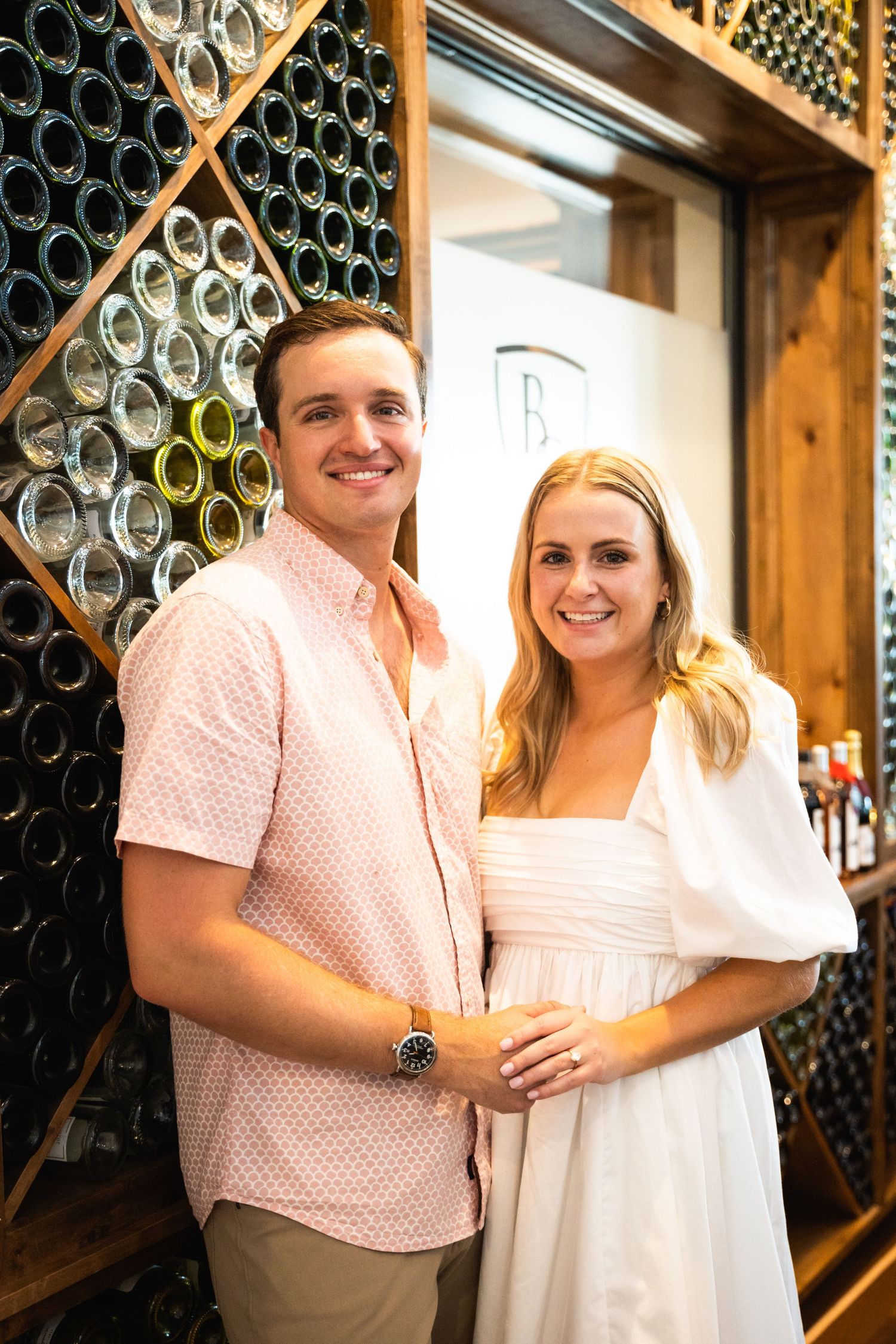 A couple poses in front of a wall of wine bottles at a winery cellar.