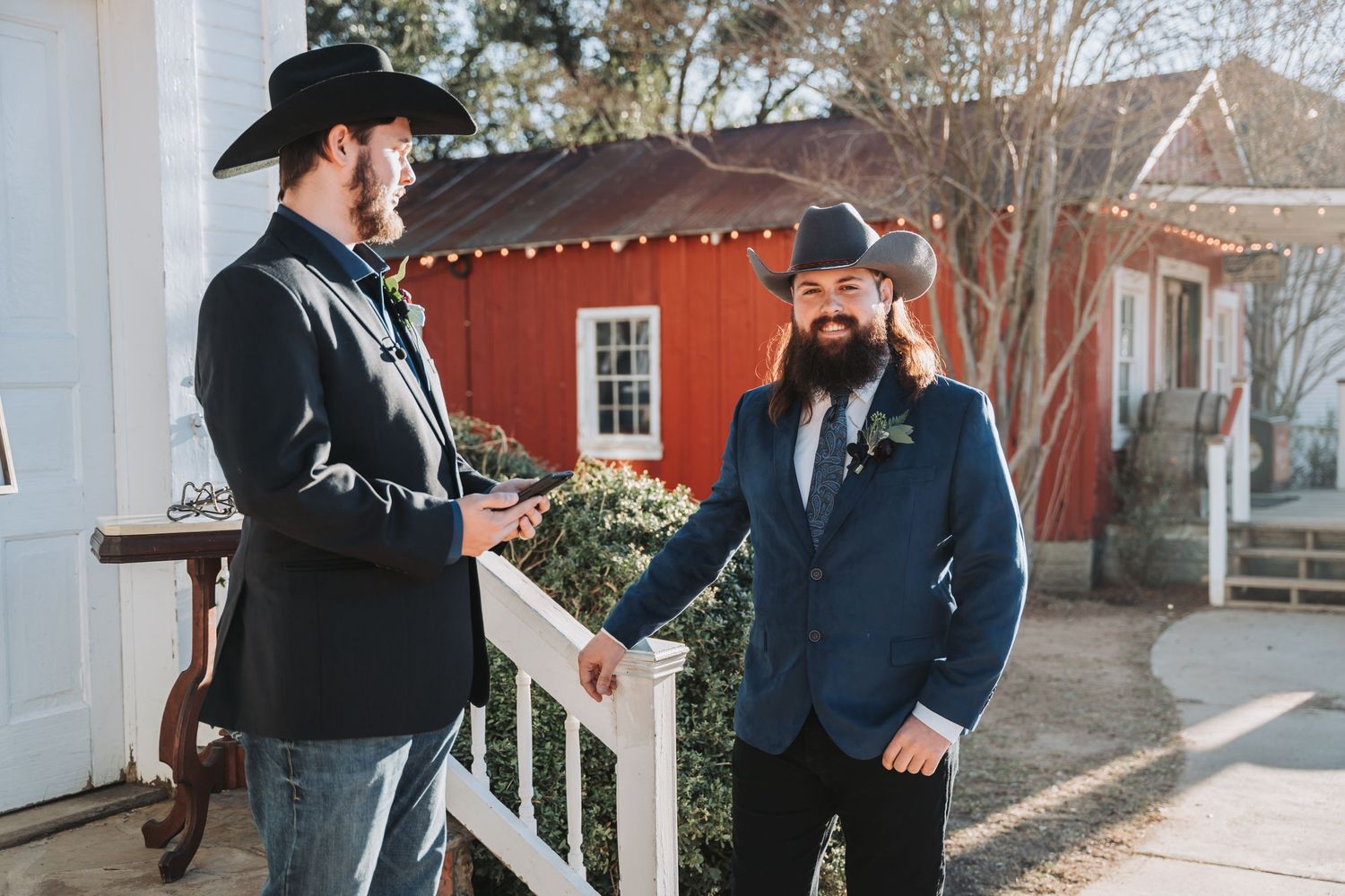 Two people in dark suits and cowboy hats chat on a porch with a red building in background.