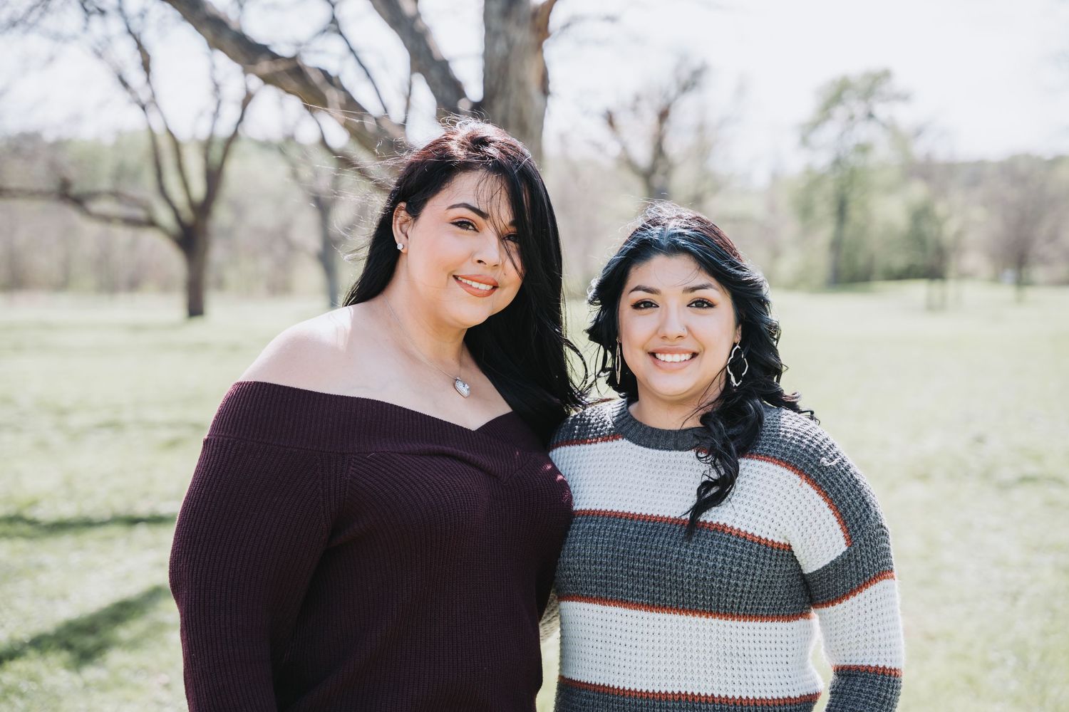 Two people in casual sweaters pose together outdoors on a sunny day with bare trees in background.