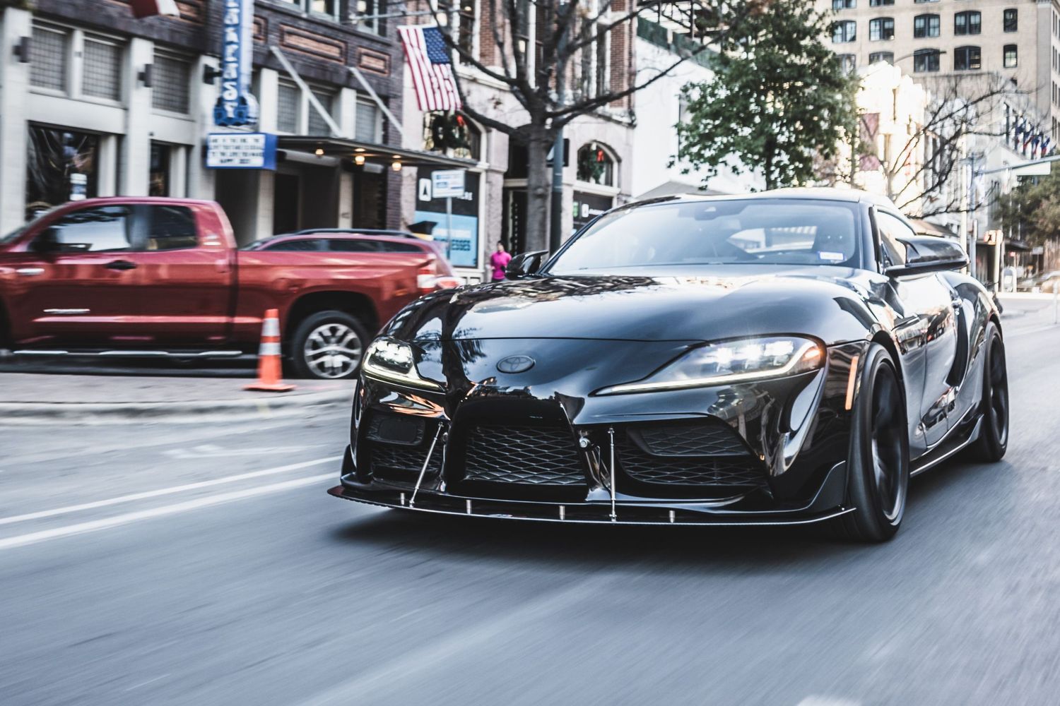Black Toyota Supra with custom body kit drives through city streets with aggressive aerodynamic modifications.