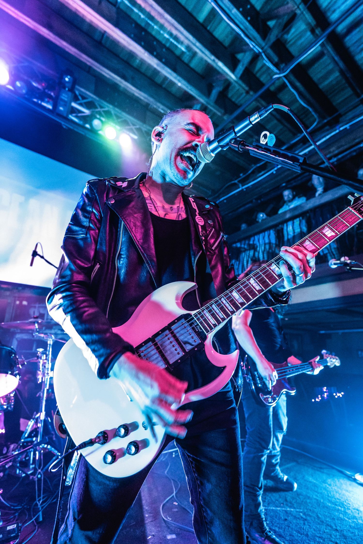 Performer in leather jacket plays white guitar under dramatic concert lighting.