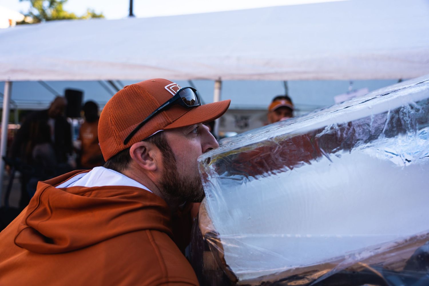 Person in orange clothing drinks from an ice sculpture at an outdoor event.