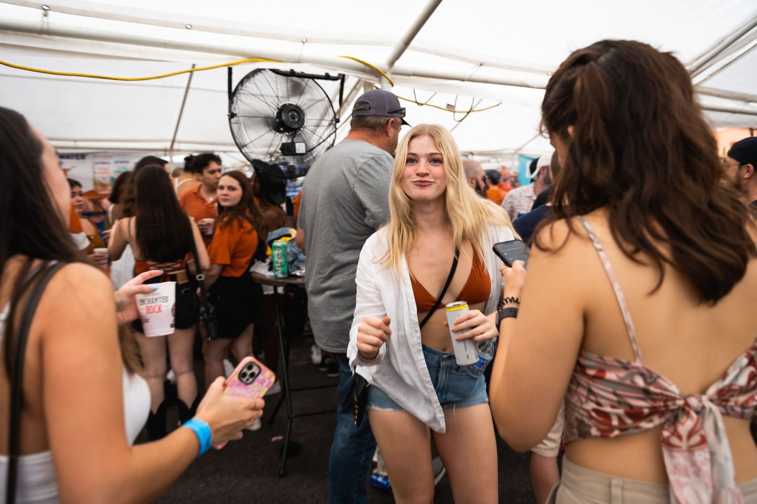 Casual crowd mingling under a white tent at a summer party with fans and lighting visible.