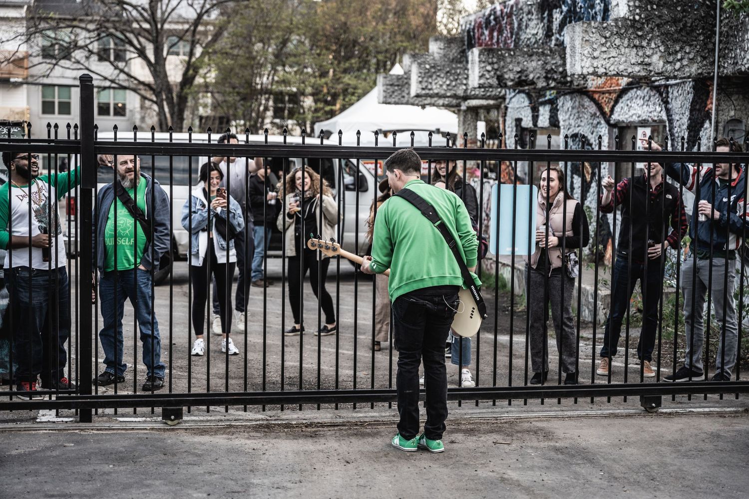 Person in green hoodie leans against metal fence at crowded outdoor event.