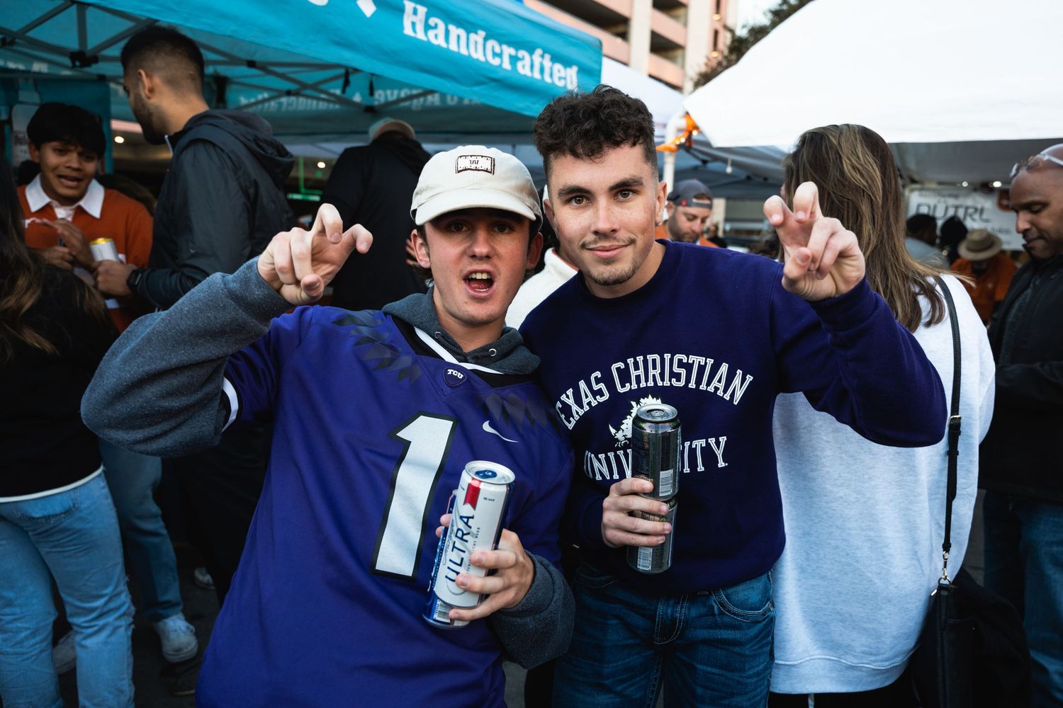 Two friends in purple team gear making peace signs while holding drinks at a tailgate.