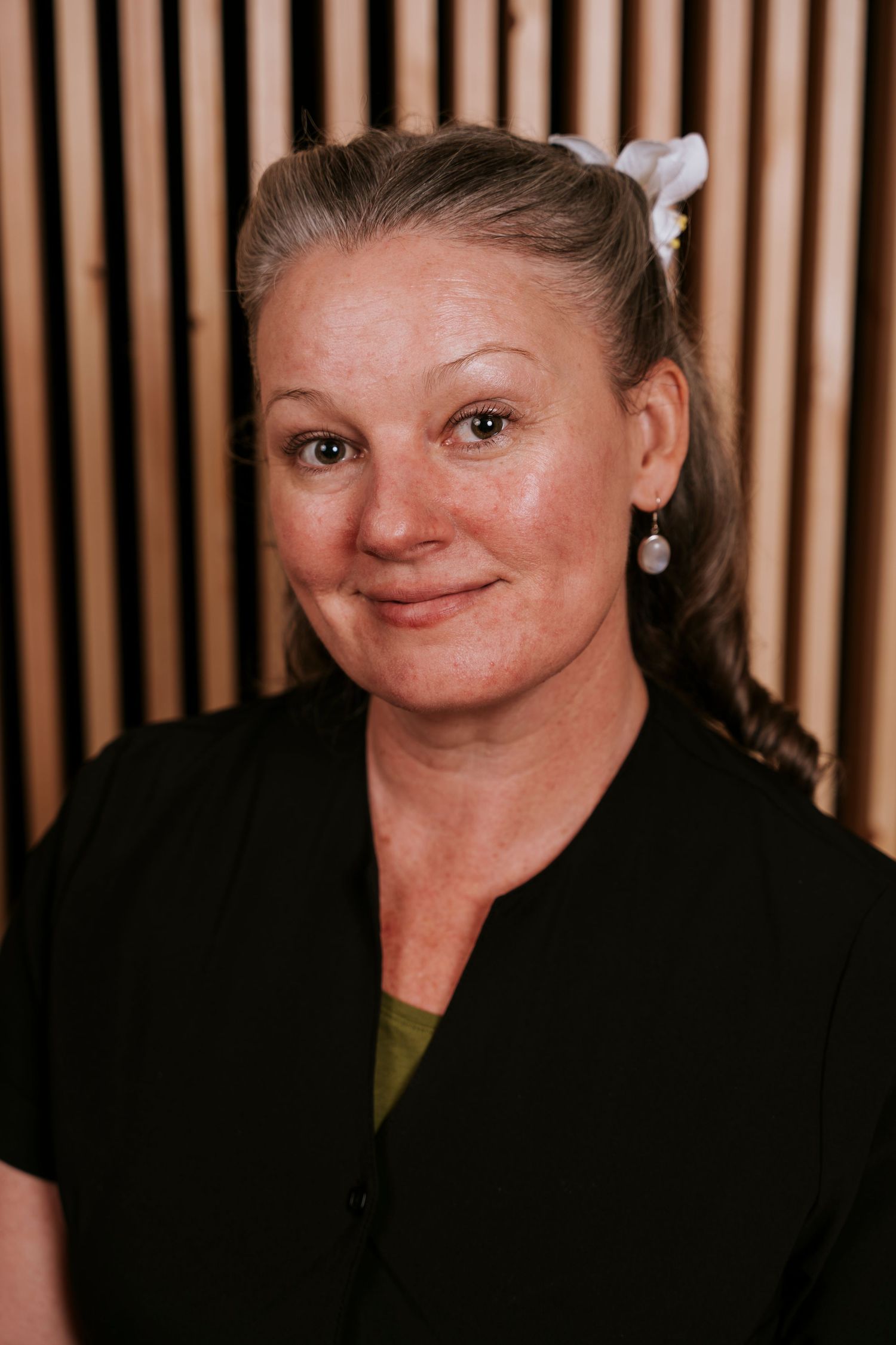 Portrait photo of someone wearing a black cardigan and olive shirt against a wood-paneled background.