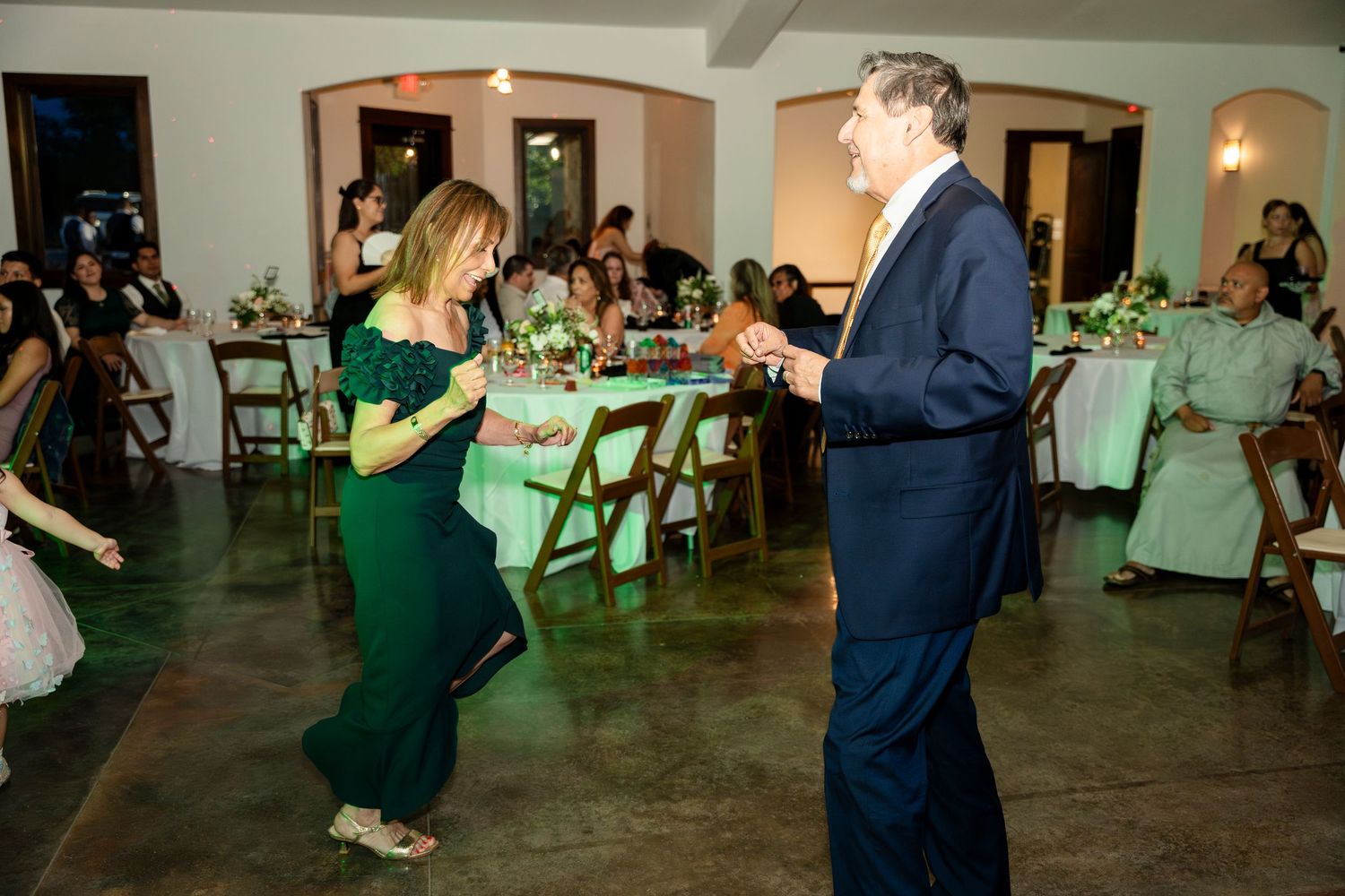A woman in green and man in navy suit dance together at an event with folding chairs and floral centerpieces visible.