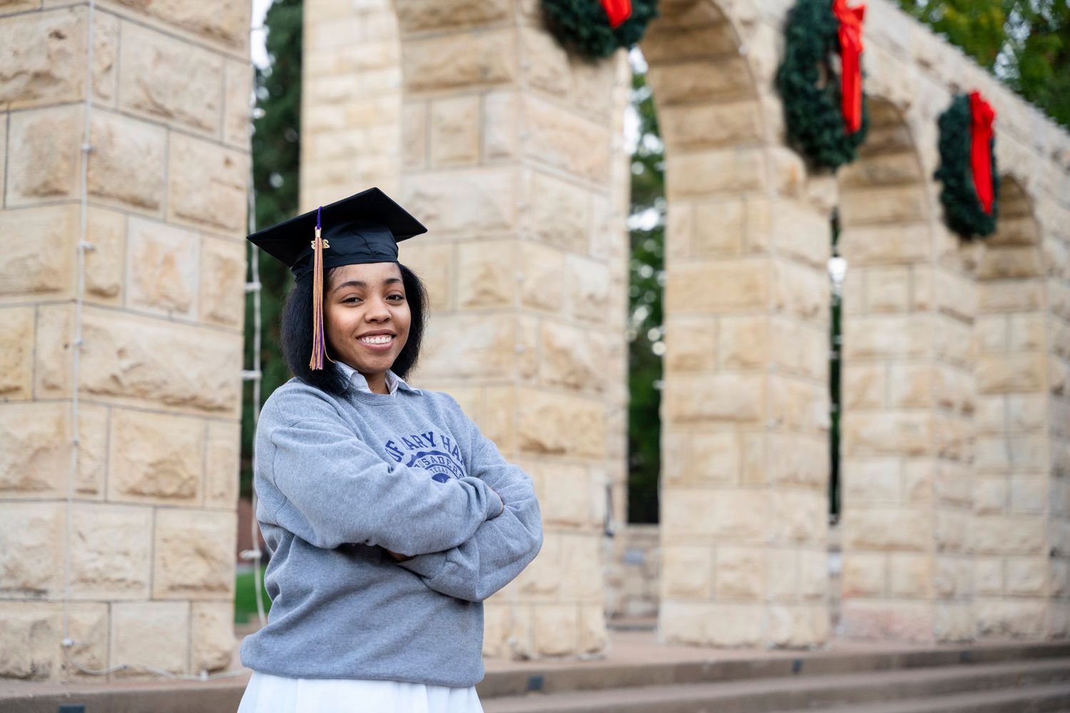 Graduate in grey university sweatshirt poses by stone archway with holiday wreaths.