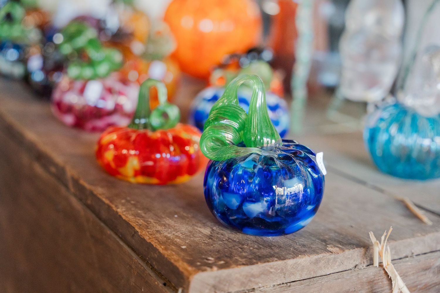 Colorful glass pumpkin decorations displayed on wooden shelf at festival market.