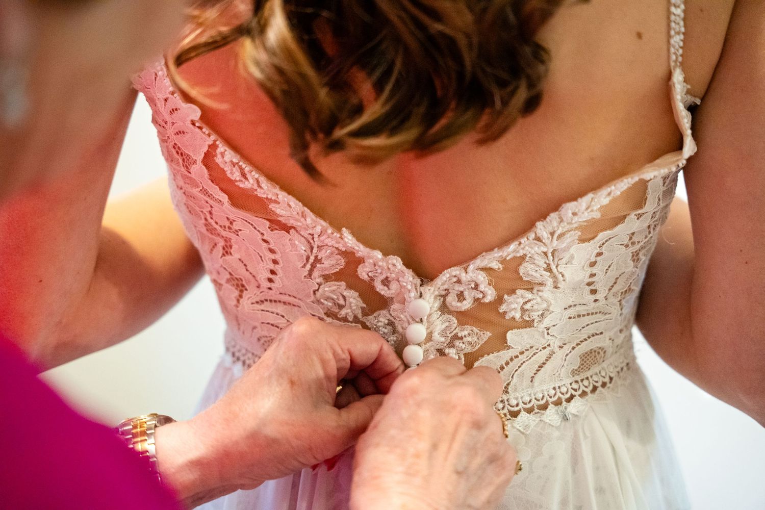 Close-up of intricate lace details being fastened on the back of a white wedding dress.