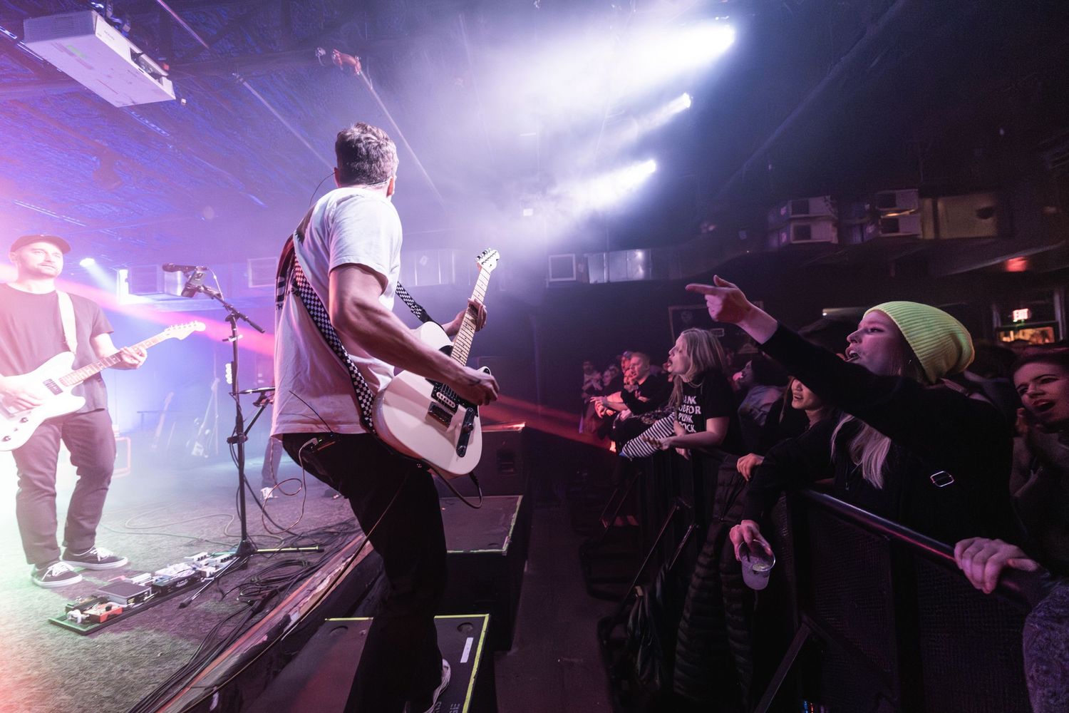 Guitarist performs at edge of stage while fans reach out from crowd barrier.