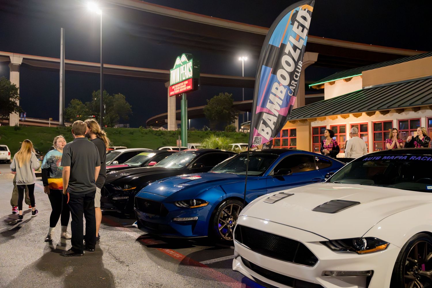 Car meet gathering at night outside a restaurant with modern Mustangs and performance vehicles.