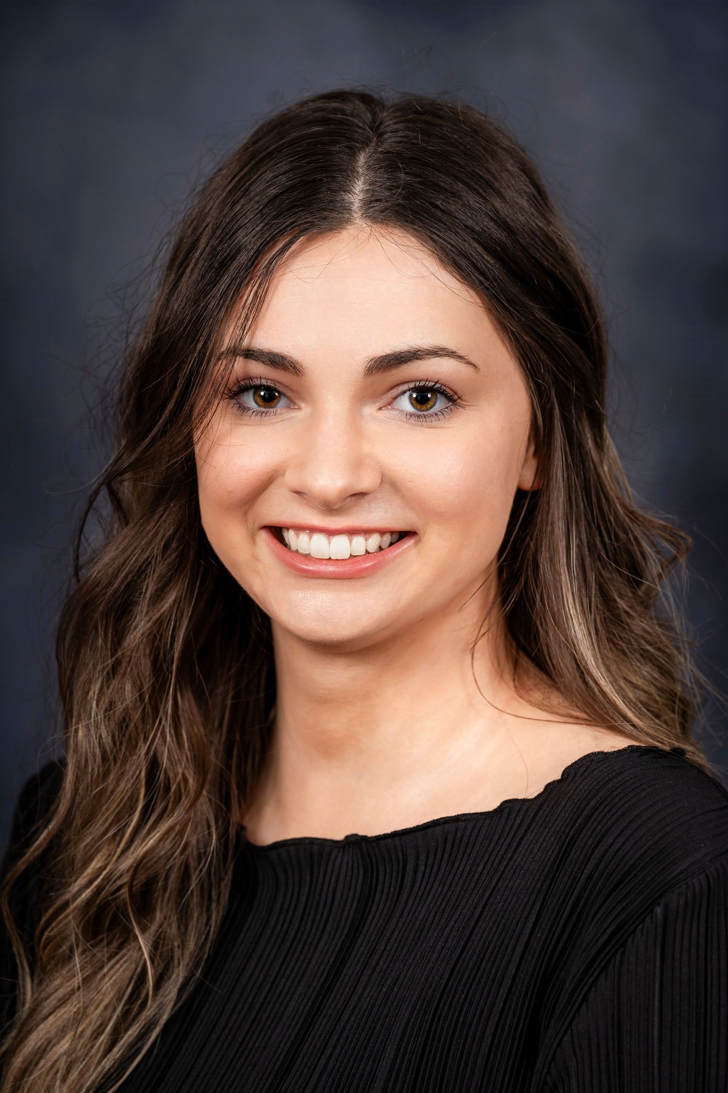 Studio portrait of a person with wavy brown hair wearing a black top and warm smile against grey backdrop.