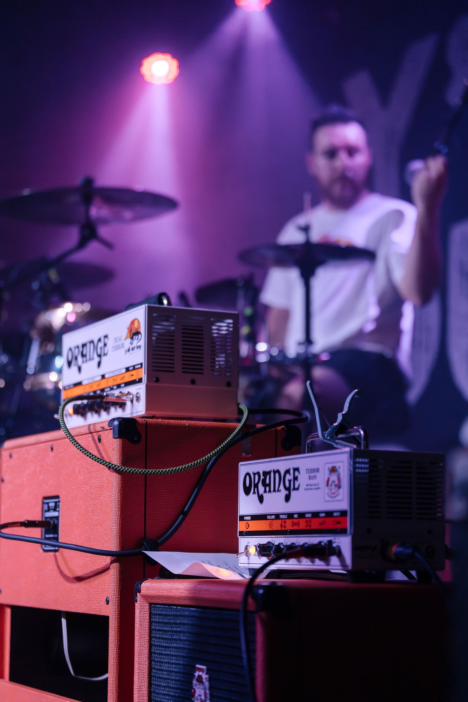 Orange amplifiers and music gear on stage with purple spotlights and drum kit in background.