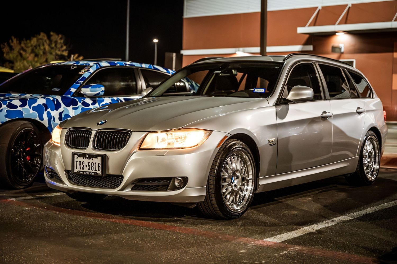 A silver BMW 3 Series wagon with custom wheels parked at night.