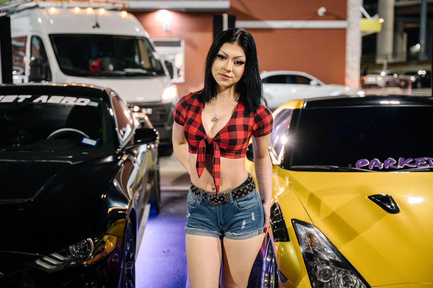 Red plaid outfit photographed between black and yellow performance cars at night.