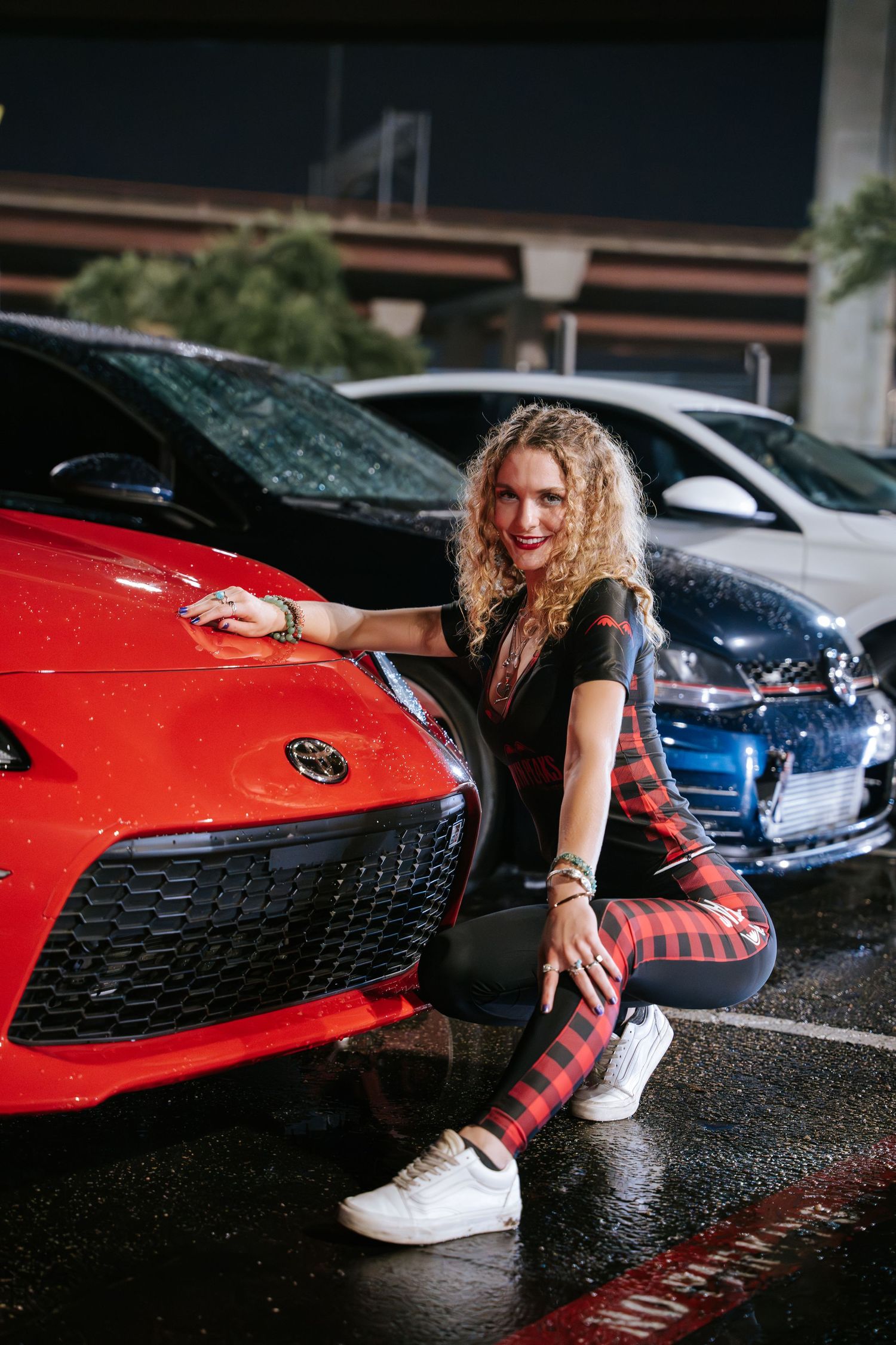 Stylish outfit pose next to bright red sports car in parking lot.