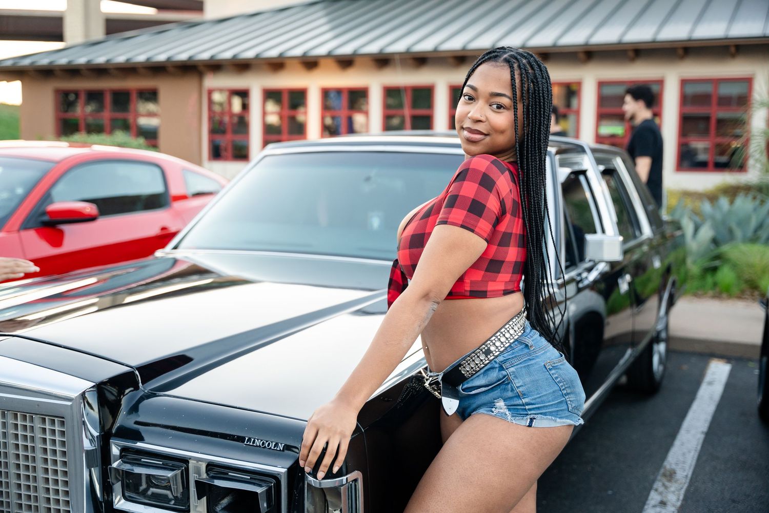 Person posing with vintage car in parking lot wearing casual attire.