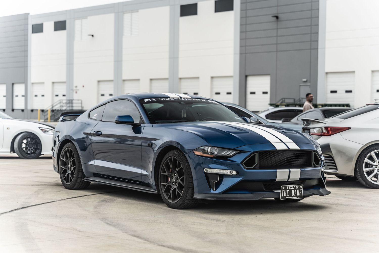 Blue Ford Mustang with racing stripes showcased in front of industrial warehouse.