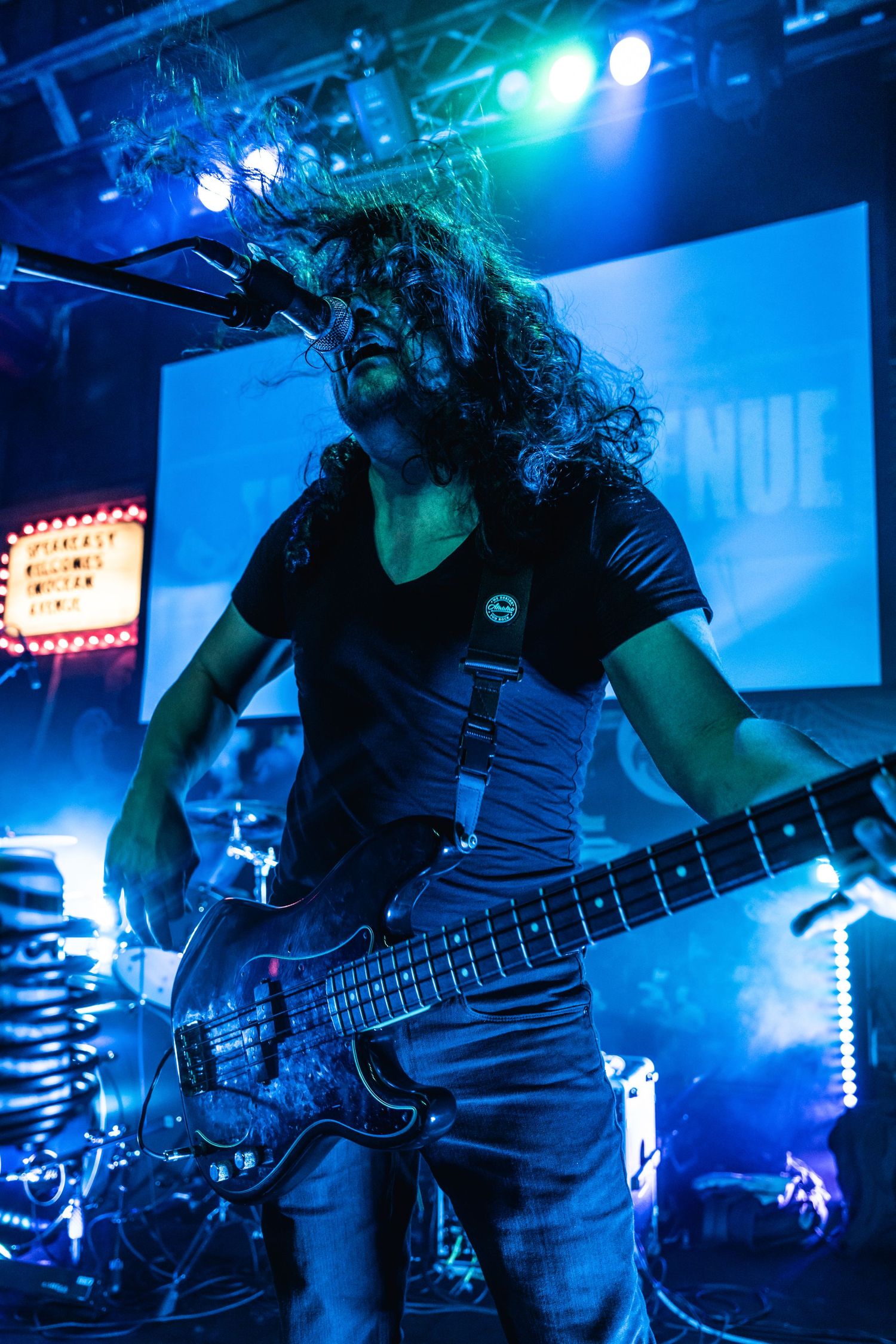 Bassist performs energetically on stage with long hair under blue concert lighting.